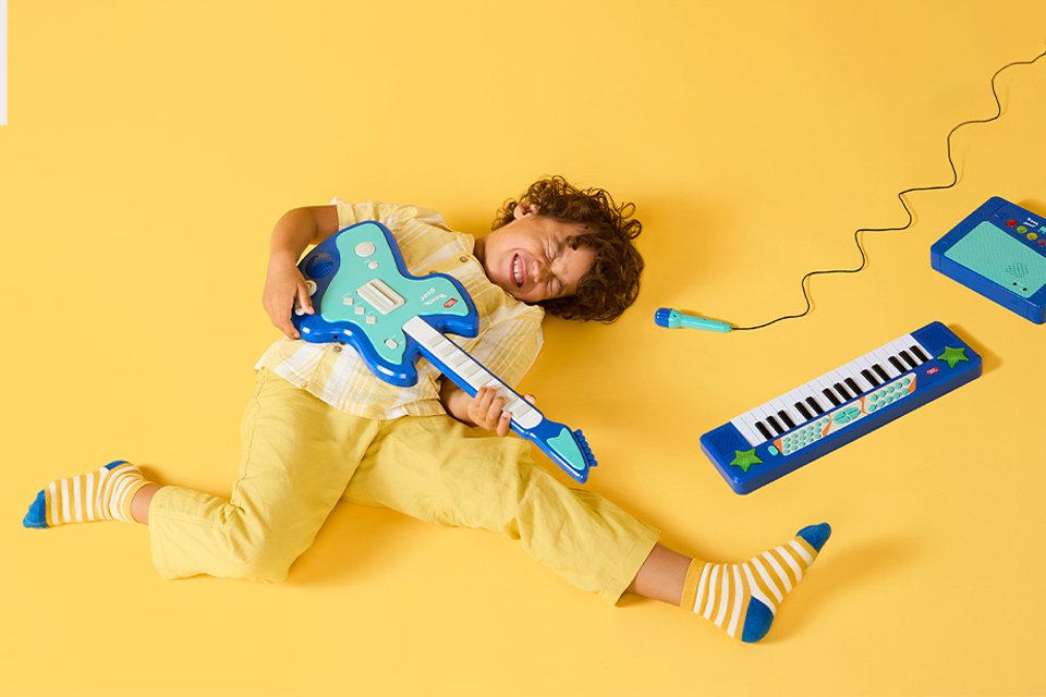 A kid holding a Chad Valley toy guitar while a keyboard placed next to him.